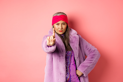 Fashion and shopping concept. Serious asian senior woman showing not so fast gesture, shaking extended finger to stop or warn you, looking determined at camera, wearing stylish purple clothes.