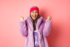 Cheerful asian senior woman winning prize and celebrating. Old funky lady saying yes and clench fists in triumph, standing over pink background.