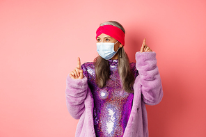 Covid-19, social distancing and fashion concept. Cheerful asian senior woman likes to party, dancing in respirator and shiny purple dress, standing over pink background.