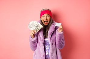 Shopping and fashion concept. Excited asian senior woman going buy something with money and plastic credit card, scream of joy and happiness, pink background.