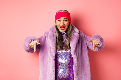 Fashion and shopping concept. Happy korean senior woman pointing fingers down and showing awesome promo deal, wearing trendy purple fur coat and headband, pink background.