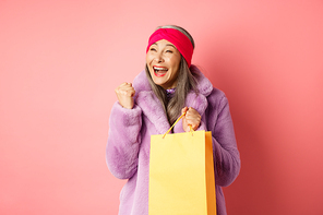Shopping and fashion concept. Happy asian senior woman winning, holding paper bag and making fist pump gesture, saying yes with cheerful face, standing over pink background.