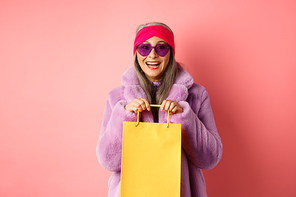 Fashionable asian senior woman going shopping in sunglasses and trendy winter clothes, holding paper bag from store and smiling happy at camera, pink background.