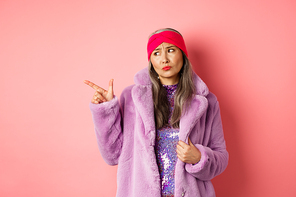 Fashion and shopping concept. Skeptical middle-aged asian woman in purple fur coat looking and pointing finger left with displeased, sad expression, standing over pink background.