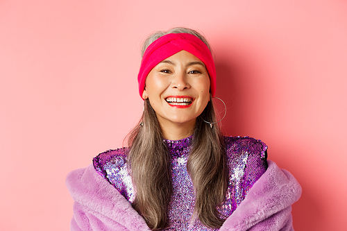 Close-up of happy asian senior lady in trendy purple outfit, laughing and smiling at camera, having fun on pink background.