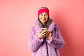 Online shopping and fashion concept. Smiling asian senior lady in winter coat and headband order in internet, holding mobile phone and looking happy, pink background.