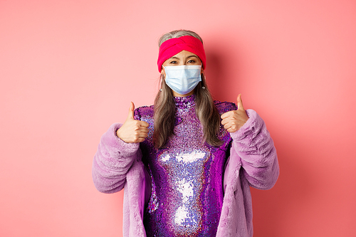 Coronavirus and shopping concept. Stylish asian grandmother in respirator and glittering purple dress and faux fur coat, showing thumbs-up, recommend shop in face masks.