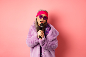 Fashion concept. Stylish senior asian woman singing karaoke, perform on stage with microphone, wearing trendy sunglasses and purple faux fur coat, pink background.