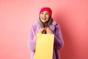 Shopping and fashion concept. Beautiful asian senior woman looking dreamy at upper left corner and imaging something, holding yellow paper bag with gift, pink background.