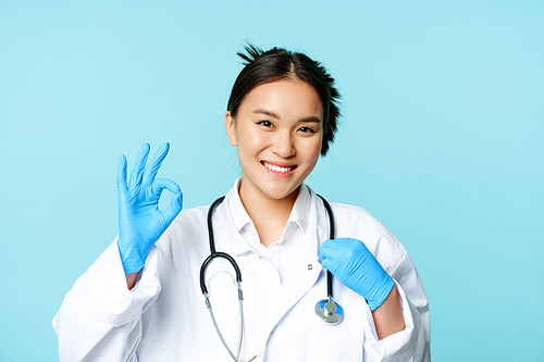 Smiling asian doctor, female physician touching stethoscope, showing okay, ok sign in approval, blue background.