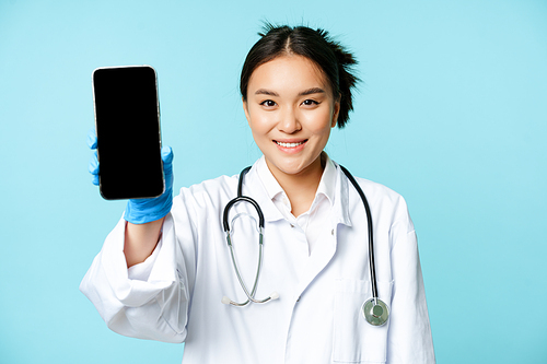 Web healthcare, online help concept. Smiling asian woman doctor, showing smartphone app interface, mobile screen, standing in uniform over blue background.