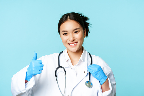 Smiling asian woman therapist, physician with stethoscope shows thumbs up, standing over blue background.
