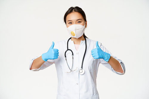 Healthcare and covid-19 quarantine concept. Smiling asian female doctor in medical respirator, showing thumbs up in sterile rubber gloves, white background.