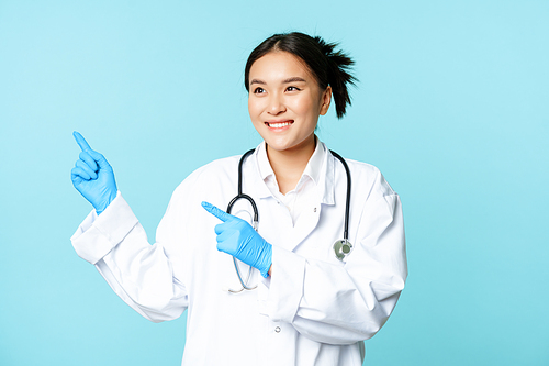 Asian smiling doctor, medical healthcare worker, pointing fingers left, looking pleased at advertisement, advertising product, blue background.