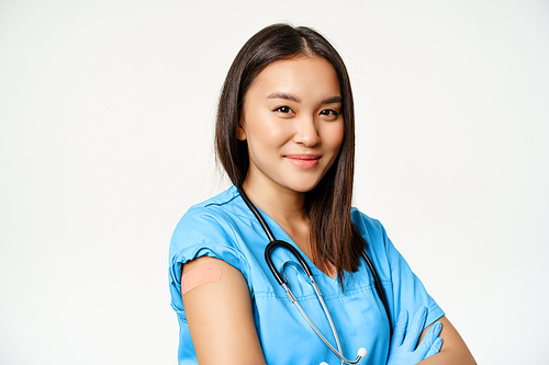 Healthcare and medical concept. Vaccinated female asian doctor showing arm with patch after covid-19 vaccine, smiling pleased, white background.