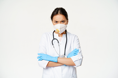 Asian woman doctor in medical face respirator and rubber gloves, looking angry at camera, cross arms on chest, standing in healthcare worker uniform, white background.