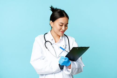 Smiling asian doctor, nurse writing down on clipboard, standing in medical uniform over blue background.