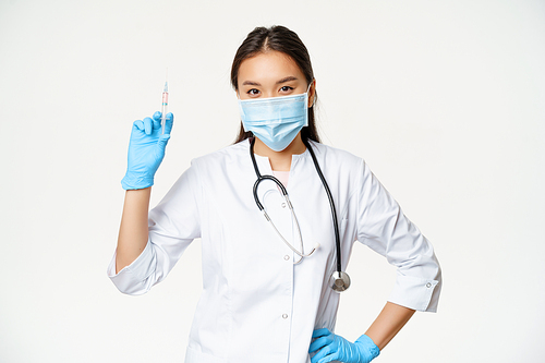 Asian nurse in medical mask holding syringe with vaccine, concept of healthcare and vaccination, standing in clinic uniform, white background.