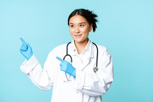 Smiling asian doctor, medical worker in uniform, pointing and looking left at copy space, standing on blue background.