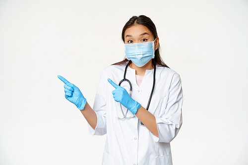 Excited asian female doctor points left, wears medical mask as covid-19 preventive measures, standing in uniform over white background.