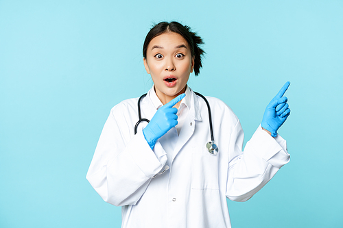 Shocked asian doctor, nurse in uniform, pointing right and gasping surprised, big news, standing over blue background.