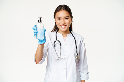 Smiling asian doctor showing hand sanitizer, bottle of antiseptic, wearing sterile rubber gloves and clinic uniform, white background.