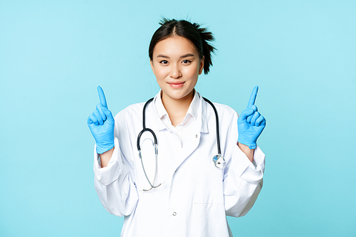 Smiling asian female doctor, nurse in medical uniform, gloves, pointing fingers up, showing healthcare information, standing over blue background.