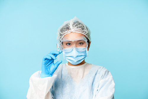 Close up portrait of asian female doctor, nurse in personal protective equipment showing syringe with vaccine, vaccinating from flu or covid, blue background.