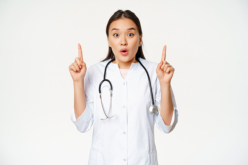 Portrait of surprised female asian doctor, physician pointing fingers up, showing amazing news, big promo, white background.