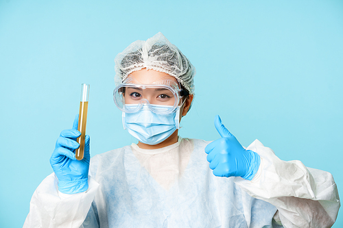 Asian female doctor, lab worker in personal protective equipment, showing thumbs up and test sample tube analise, standing over blue background.
