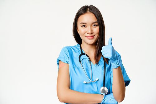 Smiling nurse, asian female doctor in scrubs, showing thumbs up sign and vaccinated arm with medical plaster, recommending vaccination from covid-19, white background.
