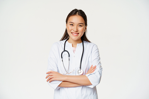 Healthcare and medical concept. Smiling female asian doctor in uniform, looking confident at camera, treating patients, standing over white background.