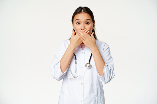 Image of shocked female asian doctor covers her lips with hands, close mouth and looking worried, taboo gesture, standing over white background.
