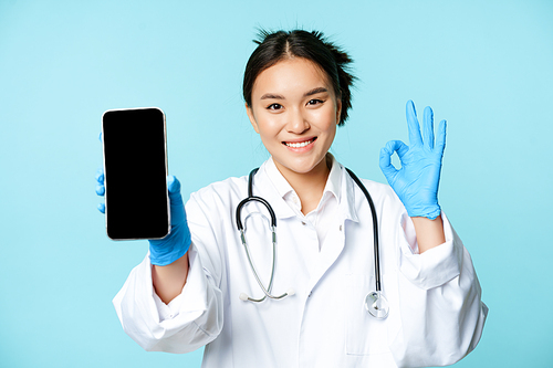 Web healthcare, online help concept. Confident asian female doctor or nurse, showing smartphone screen, app interface and okay sign in approval, recommending website.