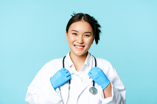 Asian woman doctor, nurse smiling confident, adjusting stethoscope on neck, standing in medical uniform over blue background.