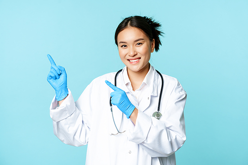 Smiling asian female doctor, therapist pointing fingers at upper left corner, showing medical advertisement, standing over blue background.
