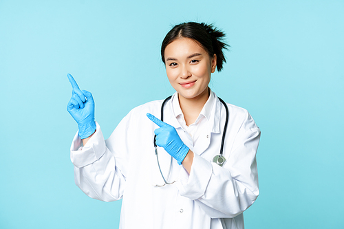 Smiling female doctor, physican in medical uniform and sterile gloves, pointing fingers left at promo, clinic logo, standing over blue background.