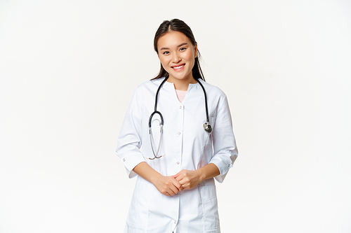 Smiling asian woman doctor, wearing medical robe and stethoscope, looking pleasant at patient, standing over white background.