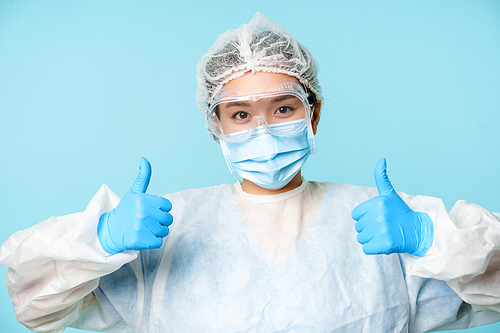 Portrait of smiling asian nurse or doctor, wearing personal protective equipment, showing thumbs up, recommending, standing over blue background.