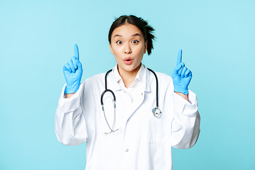 Excited asian medical worker, female doctor pointing fingers up, wearing sterile gloves and uniform, standing over blue background.
