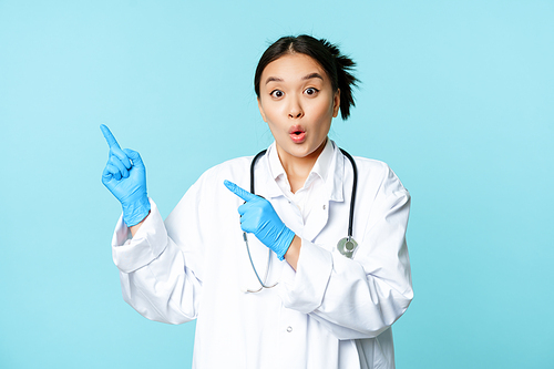 Surprised and enthusiastic asian doctor, female medical worker pointing fingers at upper left corner, showing disocunt, standing over blue background.