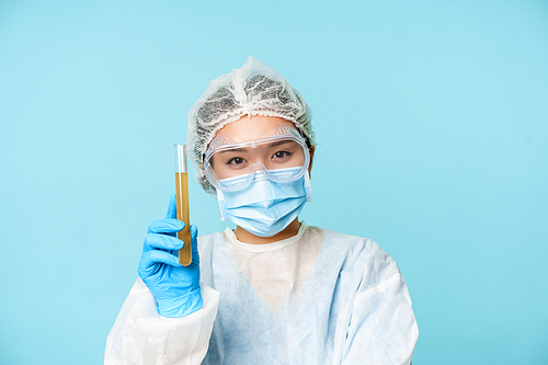 Smiling asian female doctor, nurse in personal protective equipment, showing sample test tube and smiling, making analise, standing over blue background.