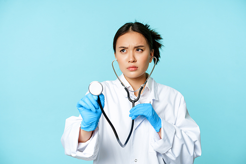 Image of serious medical worker, asian doctor listening to patient lungs with stethoscope, examining person, standing over blue background.