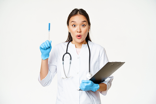 Enthusiastic nurse, asian woman doctor raising pen up, holding clipboard with patient papers, standing in uniform over white background.