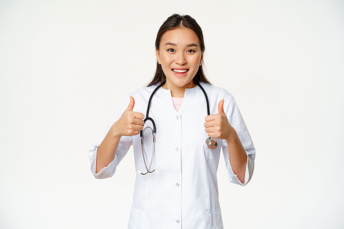 Smiling asian female doctor, wearing medical robe, showing thumbs up pleased, recommending health product, standing over white background.