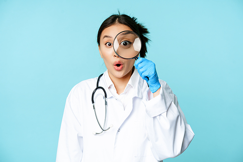 Image of asian woman doctor or nurse found smth, looking through magnifying glass and stare surprised, shocked, standing in uniform over blue background.
