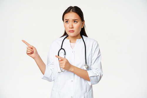 Skeptical female doctor, concerned nurse pointing and looking left with worried face expression, standing in white medical robe against studio background.