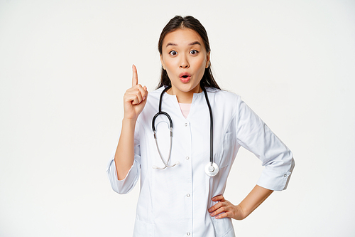 Healthcare and medical concept. Asian woman doctor raising finger, found solution, treatment for patient, pointing up and looking enthusiastic, white background.
