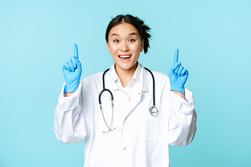 Enthusiastic asian doctor, female physician pointing fingers up and smiling, showing advertisement, demonstrating promo, blue background.
