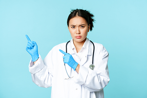 Angry female doctor or nurse, pointing fingers at upper left corner with disapproval, sulking and furrow eyebrows, standing in medical uniform, blue background.